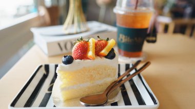 Bir dilim meyveli kek çizgili bir tabakta serinletici bir içecekle, rahat bir kafe atmosferinde.