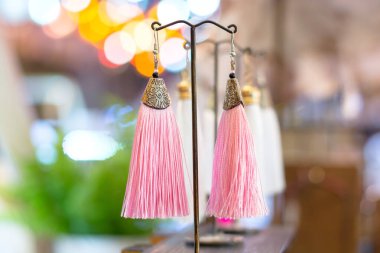 Bir stantta zarif bir şekilde sergilenen bir çift pembe püsküllü küpe, canlı renklerini ve bulanık bir arka plana karşı karmaşık tasarımlarını sergiliyor..