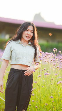 Çiçeklerin ortasında duran genç bir kadın, güneş ışığının ve doğanın tadını çıkarıyor, rahat bir hava yayan sıradan bir kıyafet giyiyor..