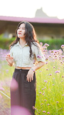 Çiçek tarlasında duran genç bir kadın, yumuşak güneş ışığıyla yıkanmış, sıradan bir kıyafet giymiş ve düşünceli bir şekilde uzaklara bakıyor..