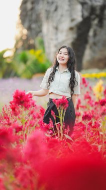 Yeşillik ve kayalık oluşumların zeminine kurulmuş canlı kırmızı çiçeklerin arasında neşeli bir kadın duruyor..