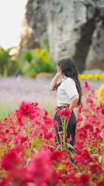 Genç bir kadın canlı kırmızı çiçeklerin arasında duruyor, doğa ile çevrili, kayalık bir zemin ile, sakin ve resimsel bir sahne yaratıyor.