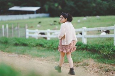 Genç bir kadın yeşil çitlerin ve arka planda otlayan hayvanlarla çevrili yeşil bir çayırda toprak bir patikada yürüyor..