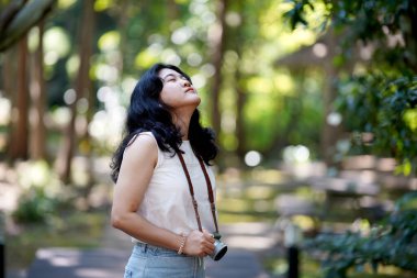 Bir kadın dışarıda duruyor, kamerayla yukarı bakıyor, yemyeşil ve yumuşak bokeh ışıklarıyla çevrili, sükuneti yakalıyor..