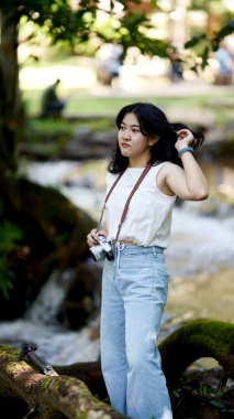 Genç bir kadın bir derenin kenarında elinde bir kamera, yeşil yapraklı ve doğal bir zemin gibi duruyor..