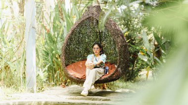 Huzurlu bir sahne, yemyeşil bir ortamda, yemyeşil bir sallanan eşsiz bir sandalyede oturan, rahatlama ve sükuneti somutlaştıran..