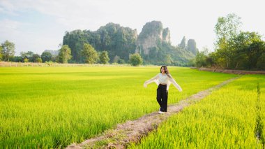 Bir kadın yemyeşil pirinç tarlasında neşeli bir şekilde duruyor, arkasında dağlar, parlak bir gökyüzünün altında..