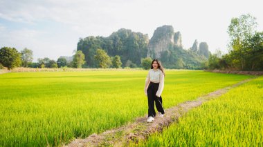 Bereketli yeşil pirinç tarlasında yürüyen bir kadın, berrak gökyüzünün altındaki yüksek dağlar tarafından çerçevelenmiş, sükuneti ve doğanın güzelliğini somutlaştıran.
