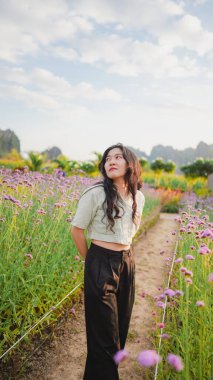 Genç bir kadın canlı bir çiçek tarlasında duruyor, yukarı bakıyor, yemyeşil ve parlak bir gökyüzünün altında açan çiçeklerle çevrili..