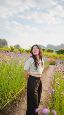 Genç bir kadın canlı bir bahçede çiçek açan çiçeklerin arasında duruyor, sakin bir ifadeyle gökyüzüne bakıyor..