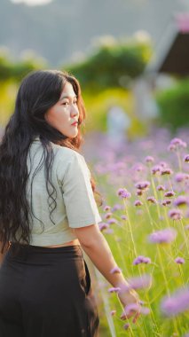 Bir kadın mor çiçeklerle dolu bir tarlada yürürken arkasına bakıyor, doğada huzurlu bir anın tadını çıkarıyor..