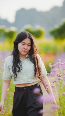 Genç bir kadın hayat dolu bir çiçek tarlasında yürüyor, doğanın güzelliğine dalmış, huzurlu bir ifadeyle..