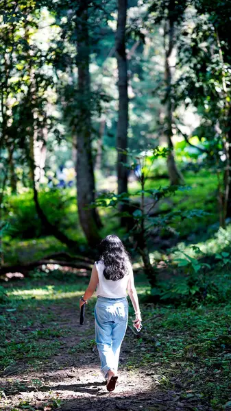 Bir kadın, yemyeşil ve uzun ağaçlarla çevrili, güneşli bir orman yolunda yürüyor, doğada huzurlu bir anın tadını çıkarıyor..