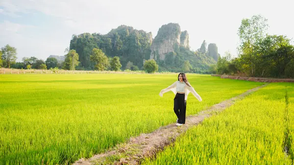 Bir kadın yemyeşil pirinç tarlasında neşeli bir şekilde duruyor, arkasında dağlar, parlak bir gökyüzünün altında..