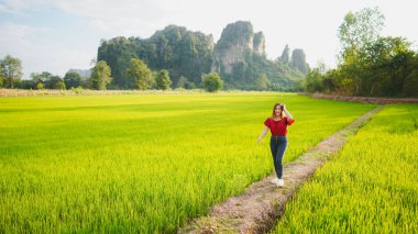 Bir kadın yemyeşil pirinç tarlasında yürüyor. Arka planda görkemli dağlar var. Güneşli güzel bir günün tadını çıkarıyor..