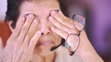 Gözlük tutarak gözlerini ovuşturan yaşlı bir adam. Yüzünde yorgunluk, stres ve rahatsızlık belirtileri var.