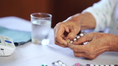 Masanın üzerinde ilaç ve kan basıncı monitörü olan su bardağı yanında beyaz hap ve su toplayan yaşlı eller.