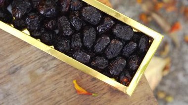 Kurumuş siyah meyve, ahşap yüzeyde altın dikdörtgen bir kutuda, arka planda bulanıklık ve yakınlarda küçük turuncu çiçek yaprağı var.