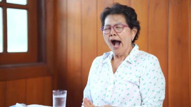 Ağzı kapalı esneyen yaşlı Asyalı kadın tarafsız bir zeminde yorgunluk ve doğal insan davranışları sergiliyor.