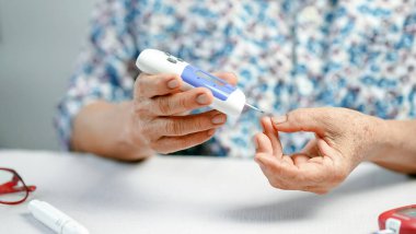 Diyabet kontrolü için kan şekeri ölçer kullanan yaşlı bir kişi, tıbbi cihazla masaya oturur, evde sağlık ve kendini izleme üzerine odaklanır