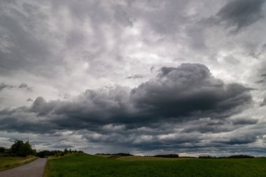 Çarpıcı fırtına bulutları kırsal bir arazide toplanıyor ve yeşil tarlalarda dolanan dar bir yol var. Sahne, yağmurdan önce gergin ve karamsar bir atmosfer yakalar.