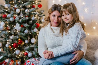 Noel arifesi beyaz süveter giyen aile kızı annesinin kucağında bir Noel ağacı ve ışıklarının arkasına yaslanır. Uzayı kopyala..