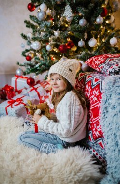 Beyaz şapkalı, uzun saçlı, elinde Noel hediyesi kutularıyla yerde oturan, beyaz kazak giyen ve Noel ağacı ve şöminenin yanındaki festival odasında poz veren mutlu küçük kız. Noel.