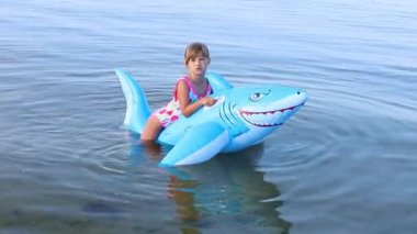 Avrupa görünümlü 7 yaşındaki mutlu kız denizde şişme köpekbalığı oyuncağında yüzerken iyi eğlenceler. Aile yaz tatili ve tatil kavramı. Yüksek kaliteli FullHD görüntüler