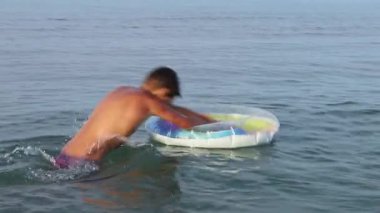Yavaş hareket eden genç, deniz suyunda şişme yüzme kucağına atlayıp dalıyor. Yaz tatilinde olan genç, suda eğleniyor. Aile tatilleri ve tatil konsepti. Yüksek kaliteli FullHD görüntüleri.