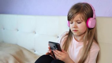 Kulaklık takan sevimli bir kız müzik dinliyor, kafasıyla dans ediyor ve evde bir yatakta oturan akıllı telefonu kullanıyor, yavaş çekimde kapatıyor. Yüksek kalite 4k görüntü