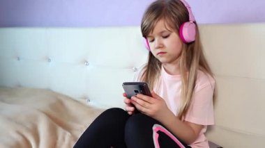 Pembe t-shirt giyen, pembe kulaklık takan, tatlı gülümseyen genç kız müzik dinleyip, akıllı telefon kullanarak dans ediyor, evde bir yatakta yavaş çekimde oturuyor. Yüksek kalite 4k görüntü