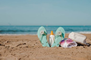 Beach essentials: flip-flops, sunscreen, towel, sunglasses, and hat on sandy shore. clipart