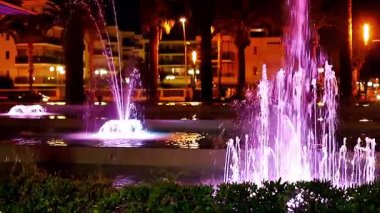 Salou, İspanya - 29 Nisan 2025: Renkli su fıskiyeleri ve parıldayan şehir manzaralı canlı gece çeşmesi. Yüksek kaliteli FullHD görüntüler