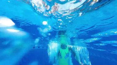 Renkli mayo giymiş genç bir kız, bereketli ağaçlar ve parlak yaz güneşinin altındaki modern binalarla çevrili, parlak bir havuza atlıyor..