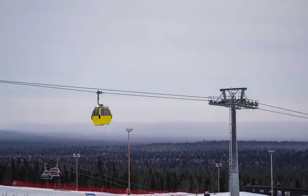 Sarı gondol karlı kış manzarasının üzerinde süzülüyor parlak sarı bir teleferik kar kaplı bir kayak merkezinin üzerinden geçiyor yoğun çam ormanları ve arka planda uzak dağlar var.. 