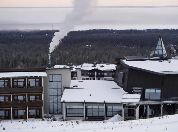 Ormanı ve dumanı olan karlı tatil köyleri Laponya 'da karlı ağaçlarla çevrili modern kış otelleri. Uzak bir bacadan yükselen duman bulutlu gökyüzüne karışıyor.. 