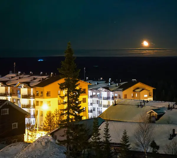 Kış oteli ay ışığında gece gökyüzünün altında karla çevrili, ılık ışıklarla parlayan rahat bir dağ beldesi ve huzurlu bir gece gökyüzü. Ay ufukta sihirli bir yansıma bırakıyor..