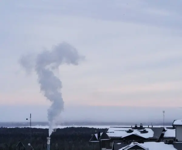 Fabrika bacasından yükselen dumanlar yoğun endüstriyel dumanlar karlı bir köyün üstündeki gökyüzüne doğru sürükleniyor, sakin kış manzarası ve uzak ufuklarla çelişiyor..