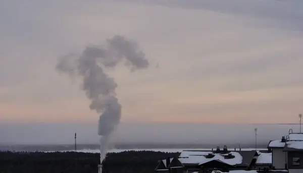 Fabrika dumanı karlı kış manzarası üzerinde sürükleniyor endüstriyel bacadan yoğun bir duman bulutu yükseliyor ve sakin, karla kaplı bir kasabanın üstündeki soğuk akşam gökyüzüne yayılıyor..