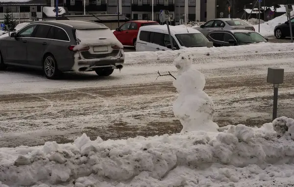 Kirli kardan adam kış otoparkında tek başına duruyor eriyen, kolları sopalı kardan adam, karlı bir otoparkta hafifçe eğiliyor, buzlu, dengesiz kar yığınlarıyla çevrili..