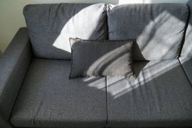 Dikdörtgen yastıklı minimalist gri kumaş kanepe, pencere perdelerinden geçen doğal ışıkla aydınlatılmış, kapalı bir alanda yumuşak gölgeler bırakılmış..