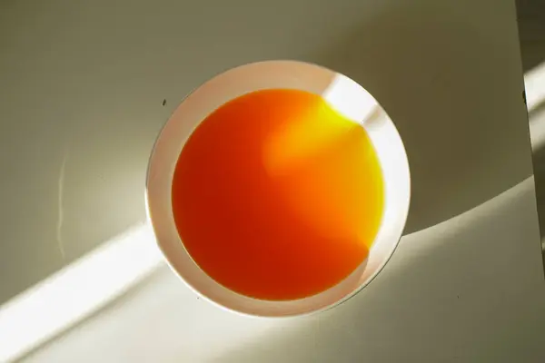 Yuvarlak turuncu ve sarı jöleli beyaz bir kasenin minimalist üst görüntüsü, temiz bir yüzeyde yumuşak gölgeler ve ılık doğal ışıkla aydınlatılıyor..