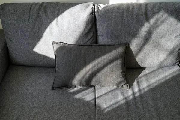 Modern gri kanepede dinlenen zarif dikdörtgen yastık, jaluziler arasından yumuşak güneş ışığıyla aydınlatılıyor, huzurlu bir iç mekanda rahatlatıcı gölgeler yaratıyor..