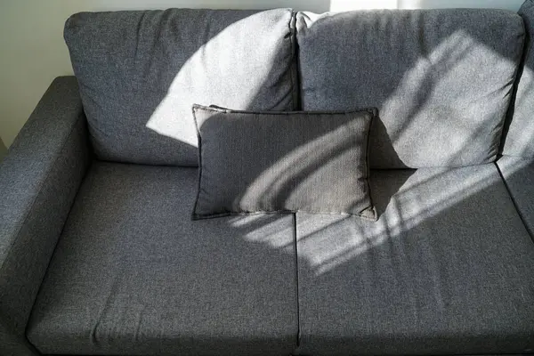 Dikdörtgen yastıklı minimalist gri kumaş kanepe, pencere perdelerinden geçen doğal ışıkla aydınlatılmış, kapalı bir alanda yumuşak gölgeler bırakılmış..