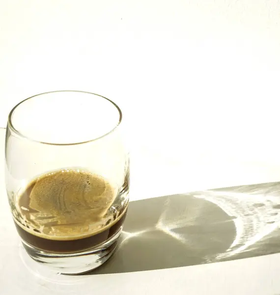 Yeni demlenmiş espresso ile berrak bir cam beyaz bir yüzeyde oturur, güçlü güneş ışığını yakalar ve minimalist bir görünüm için dramatik gölgeler ve yansımalar yaratır..
