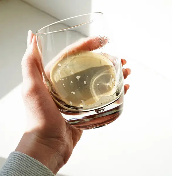 Kadın eli, parlak güneş ışığında ferahlatıcı bir limon içeceğiyle şeffaf bir bardak tutuyor. Temiz çizgiler ve yumuşak gölgelerle minimalist kompozisyon.