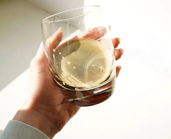 Kadın eli, şeffaf bir bardağı taze espresso ile tutar. Sabah güneşiyle aydınlatılır. Sıcak tonlu ve keskin gölgeli minimalist kompozisyon.