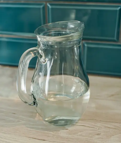 Yarı dolu şeffaf bir sürahi hafif ahşap bir tezgahın üzerine yerleştirilir. Sürahi kolu sola dönük, seramik seramik seramik seramik bir zemin üzerinde duruyor. 
