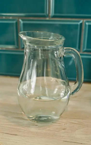 Temiz bir cam sürahi, arka planda turkuaz fayanslar bulunan ahşap bir tezgahın üzerinde duruyor. Sürahi tamamen boş, rahat bir mutfak ortamında yumuşak yansımaları yakalıyor..