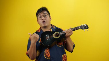 Geleneksel Hawaii gömleği giyen Asyalı genç şarkıcı sarı arka planda akustik gitar tutuyor.